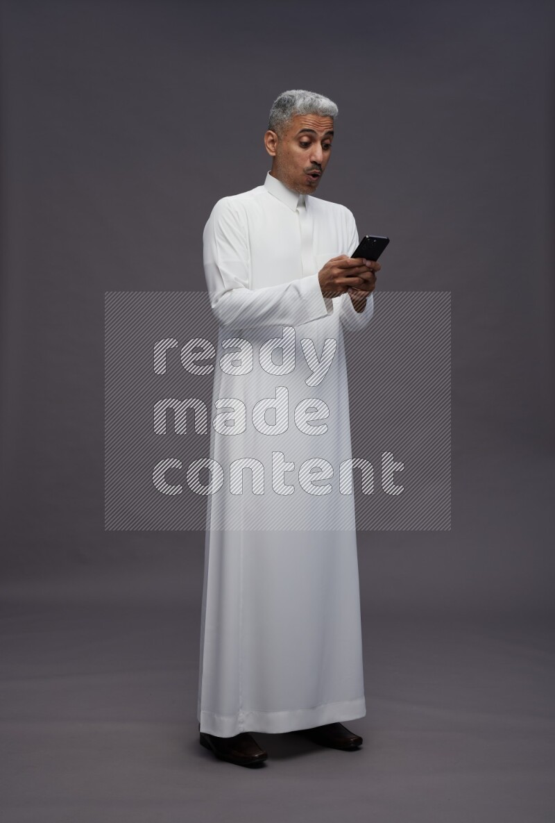 Saudi man wearing thob standing texting on phone on gray background