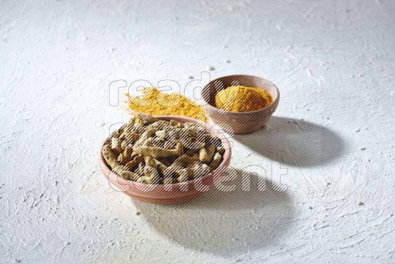 وعاءان خشبيان، أحدهما ممتلئ ببودرة الكركم والآخر بأصابع الكركم المجففة، على خلفية بيضاء