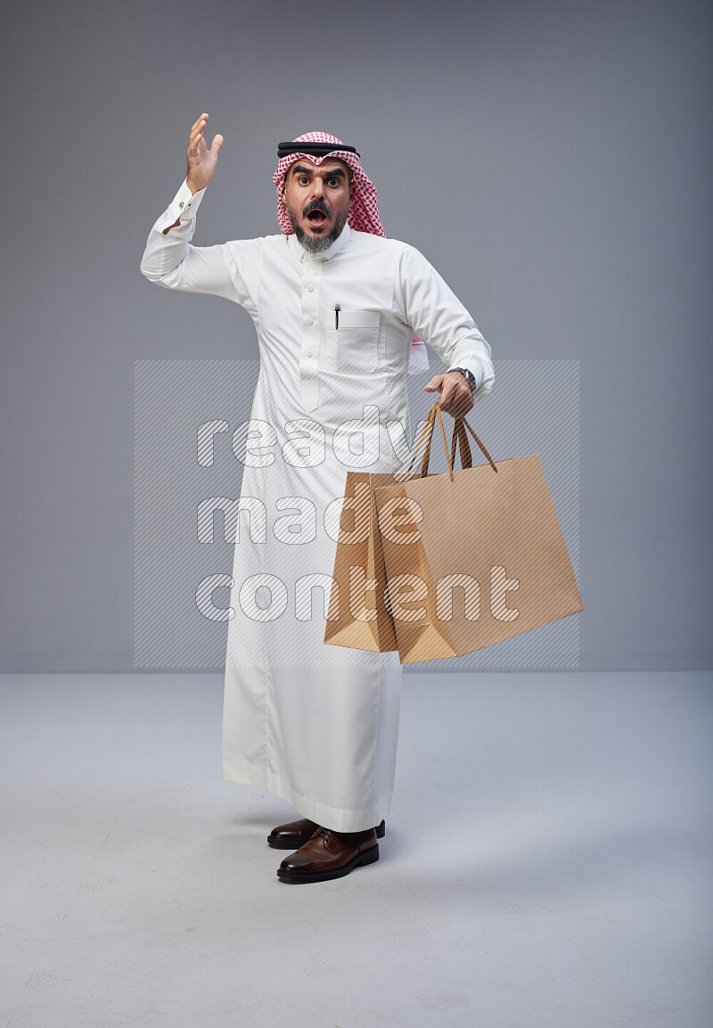 Saudi man Wearing Thob and red Shomag standing holding shopping bag on Gray background