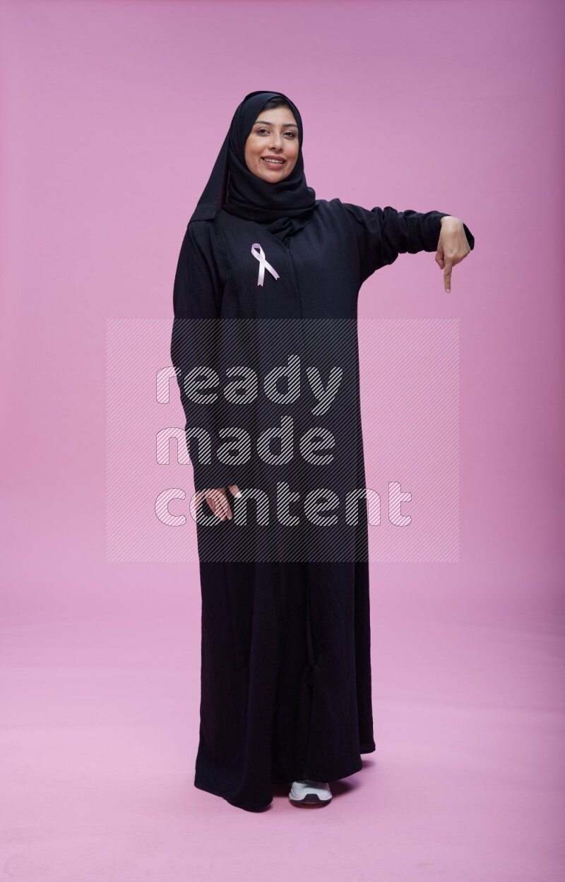 Saudi woman wearing pink ribbon on Abaya standing interacting with the camera on pink background