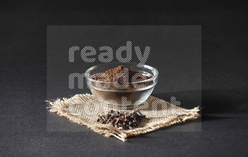 A glass bowl full of cloves powder with cloves grains on a burlap piece on a black flooring