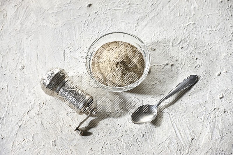 A glass bowl full of white pepper powder with white pepper beads and a metal grinder on textured white flooring