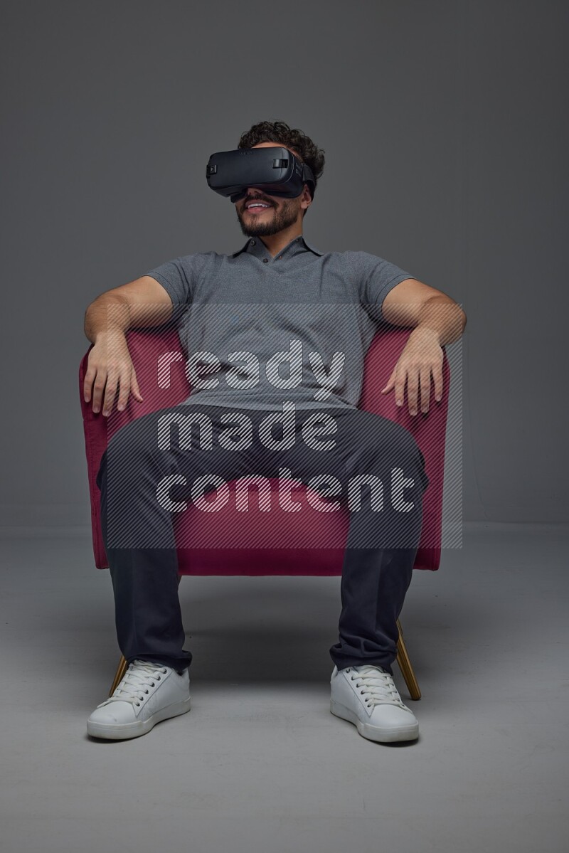 A man wearing casual and wearing VR while sitting on a burgundy chair eye level on a gray background
