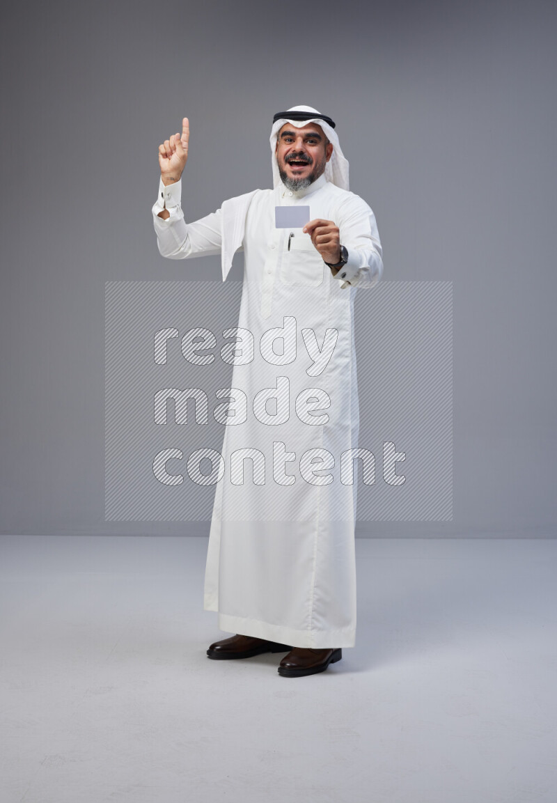 Saudi man Wearing Thob and white Shomag standing holding ATM card on Gray background