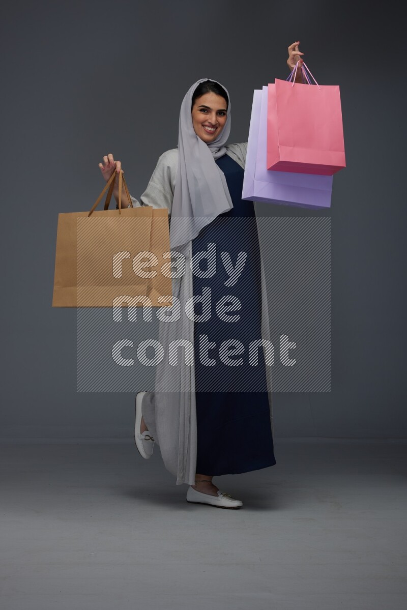 A Saudi woman wearing a light gray Abaya and head scarf standing and holding shopping bags on a grey background