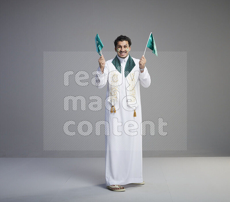 A Saudi man standing wearing thob and saudi flag scarf with face painting holding small Saudi flag on gray background