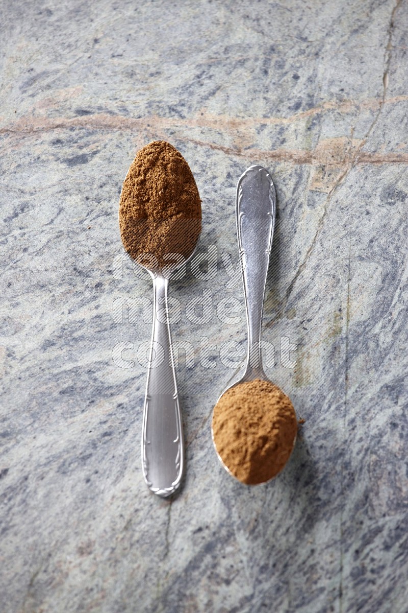 Two metal spoons full of cinnamon powder on grey marble background