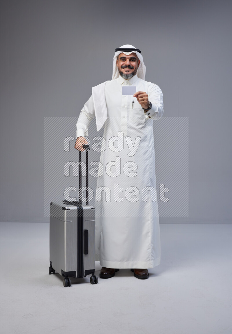 Saudi man wearing Thob and white Shomag standing holding Travel bag and ATM card on Gray background