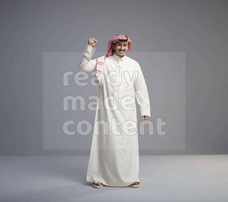 A Saudi man standing wearing thob and red shomag interacting with the camera on gray background