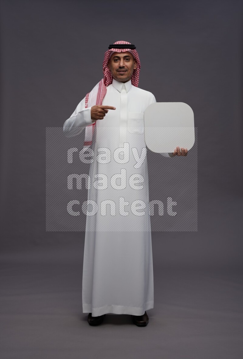 Saudi man wearing thob and shomag standing holding social media sign on gray background