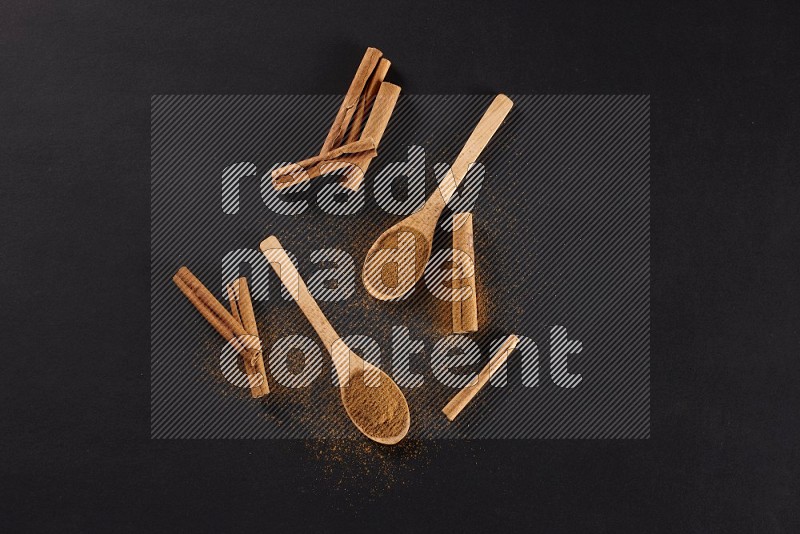 Cinnamon powder in two wooden spoons with cinnamon sticks on black background