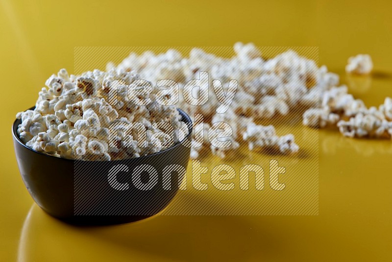 A black ceramic bowl full of popcorn with popcorn beside it on a yellow background in different angles