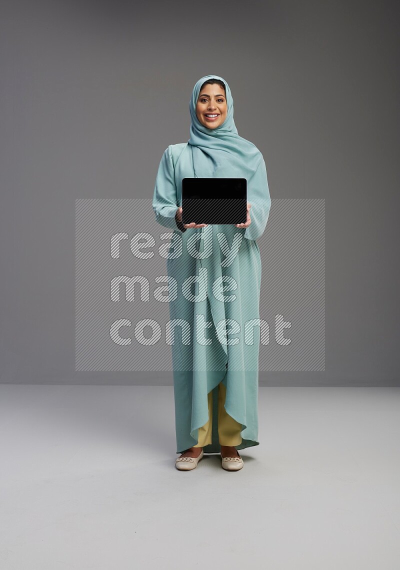 Saudi Woman wearing Abaya standing showing tablet to camera on Gray background