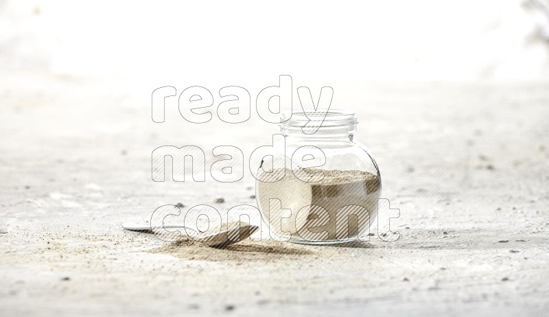 وعاء زجاجي عشبي ممتلئ ببودرة الفلفل الأبيض وملعقة معدنية ممتلئة بالبودرة على أرضية بيضاء
