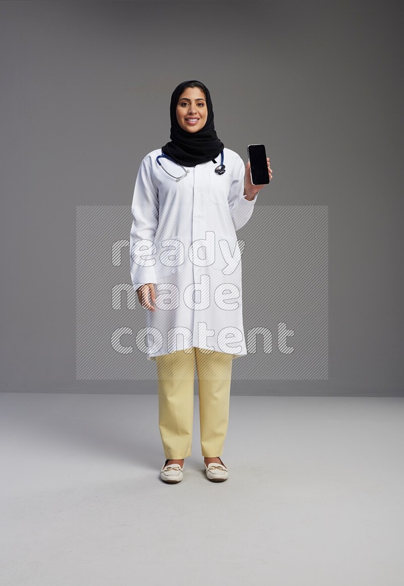 Saudi woman wearing lab coat with stethoscope standing showing phone to camera with sign in the back on Gray background