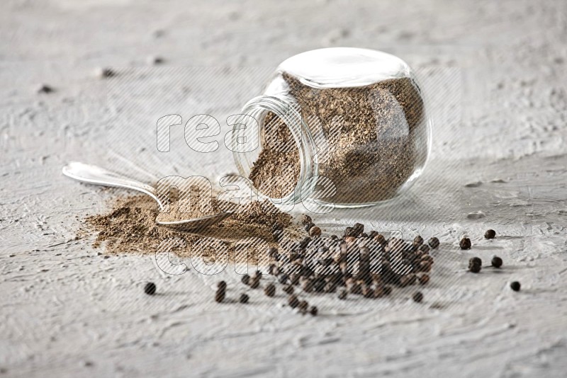 Flipped glass spice jar full of black pepper powder with a metal spoon and black pepper beads spread on a textured white flooring