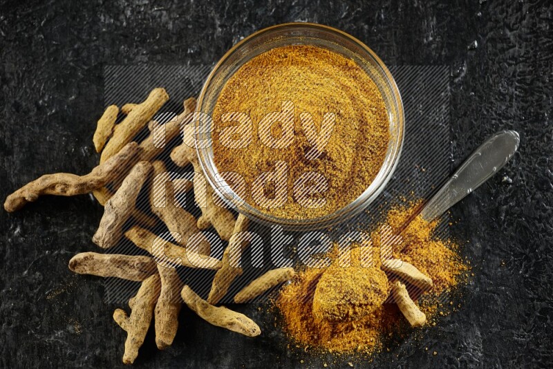 A glass bowl and a metal spoon full of turmeric powder and dried whole fingers next of them on textured black flooring