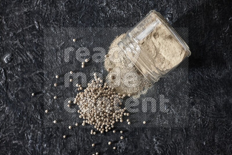 A flipped glass jar full of white pepper powder with spilled powder and pepper beads on textured black flooring