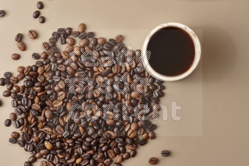 A beige pottery cup of coffee surrounded by roasted coffee beans on beige background