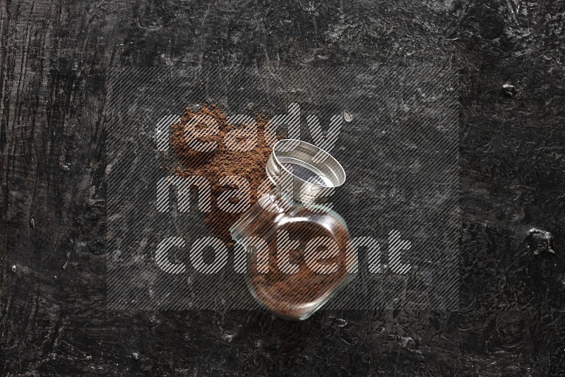 A flipped glass spice jar full of cloves powder and powder came out of it on textured black flooring