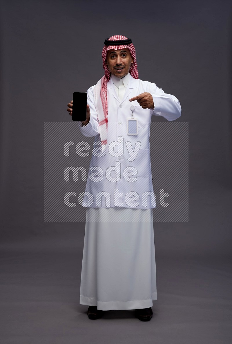 Saudi man wearing thob with lab coat and shomag with pocket employee badge standing showing phone to camera on gray background