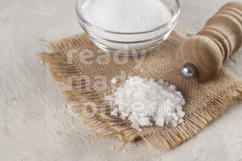 A glass bowl full of white salt with a wooden grinder on a burlap fabric all on white background