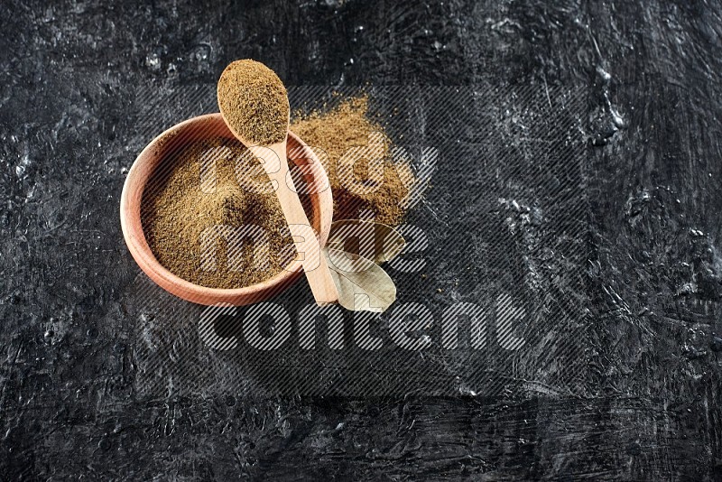 A wooden bowl and spoon full of cumin powder on a textured black flooring