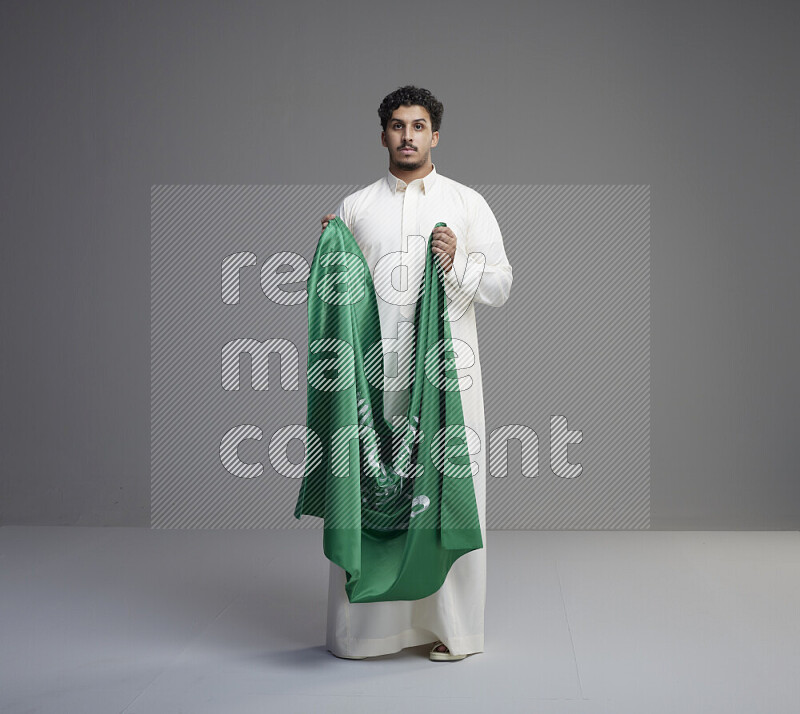 A saudi man standing wearing thob holding big saudi flag on gray background