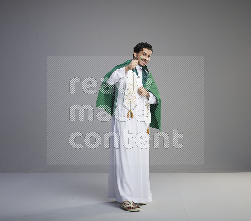 A Saudi man standing wearing thob and saudi flag scarf with face painting wrapping big Saudi flag on gray background