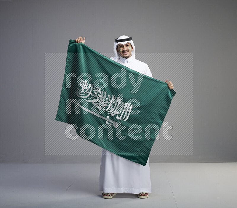 A Saudi man standing wearing thob and white shomag with face painting holding big Saudi flag on gray background