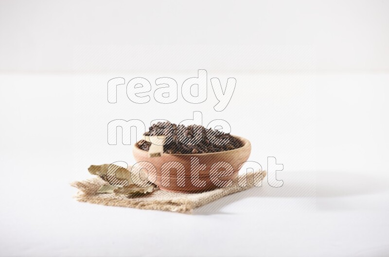 A wooden bowl and a wooden spoon full of cloves on a piece of burlap on a white flooring