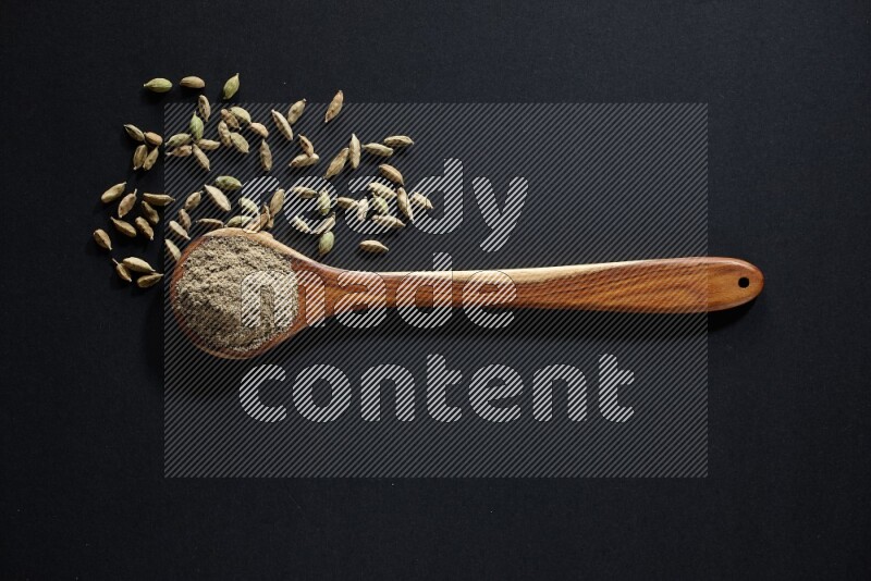 A wooden ladle full of cardamom powder and cardamom seeds beside it on black flooring