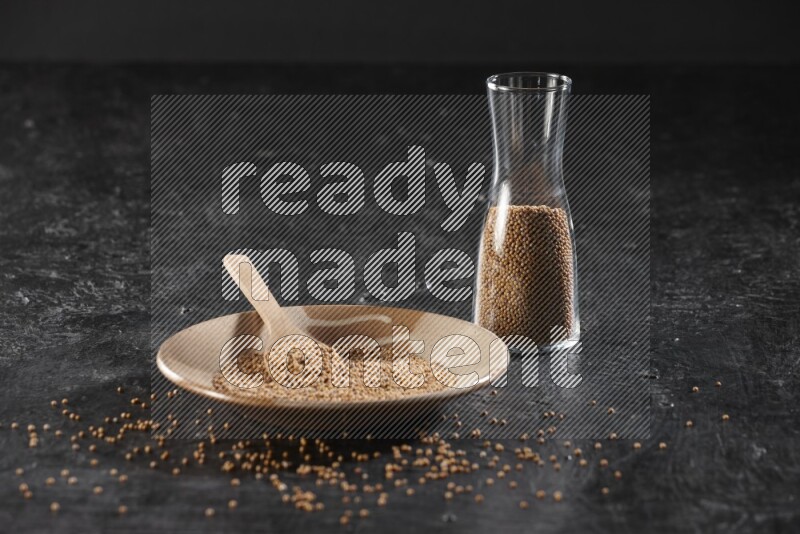 A beige pottery plate full of mustard seeds and a wooden spoon in it with a glass jar filled with the seeds on a textured black flooring