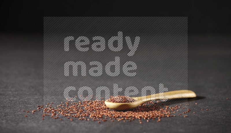 A wooden spoon full of garden cress seeds and seeds spread beside it on a black flooring