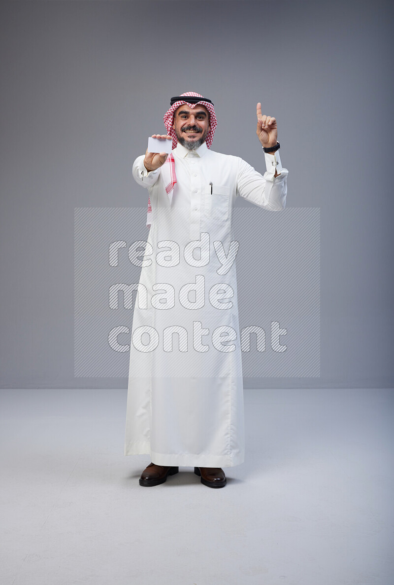 Saudi man Wearing Thob and red Shomag standing holding ATM card on Gray background