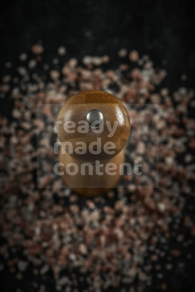 A wooden grinder standing upright and surrounded by coarse pink himalayan salt on black background