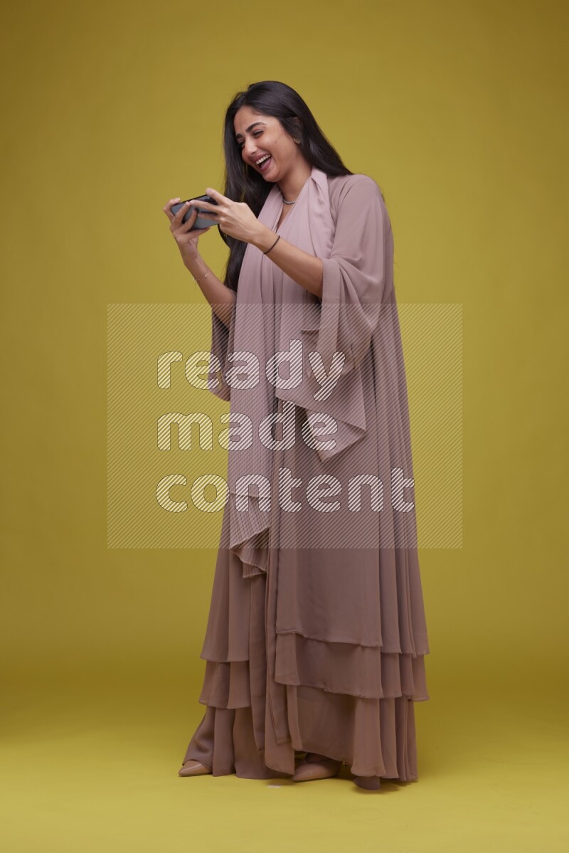 A woman Playing a Game on her smartphone on a Yellow Background wearing Brown Abaya