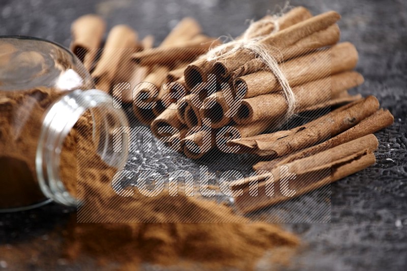 Herbal glass jar full of cinnamon powder flipped with cinnamon sticks stacked and bounded on a textured black background
