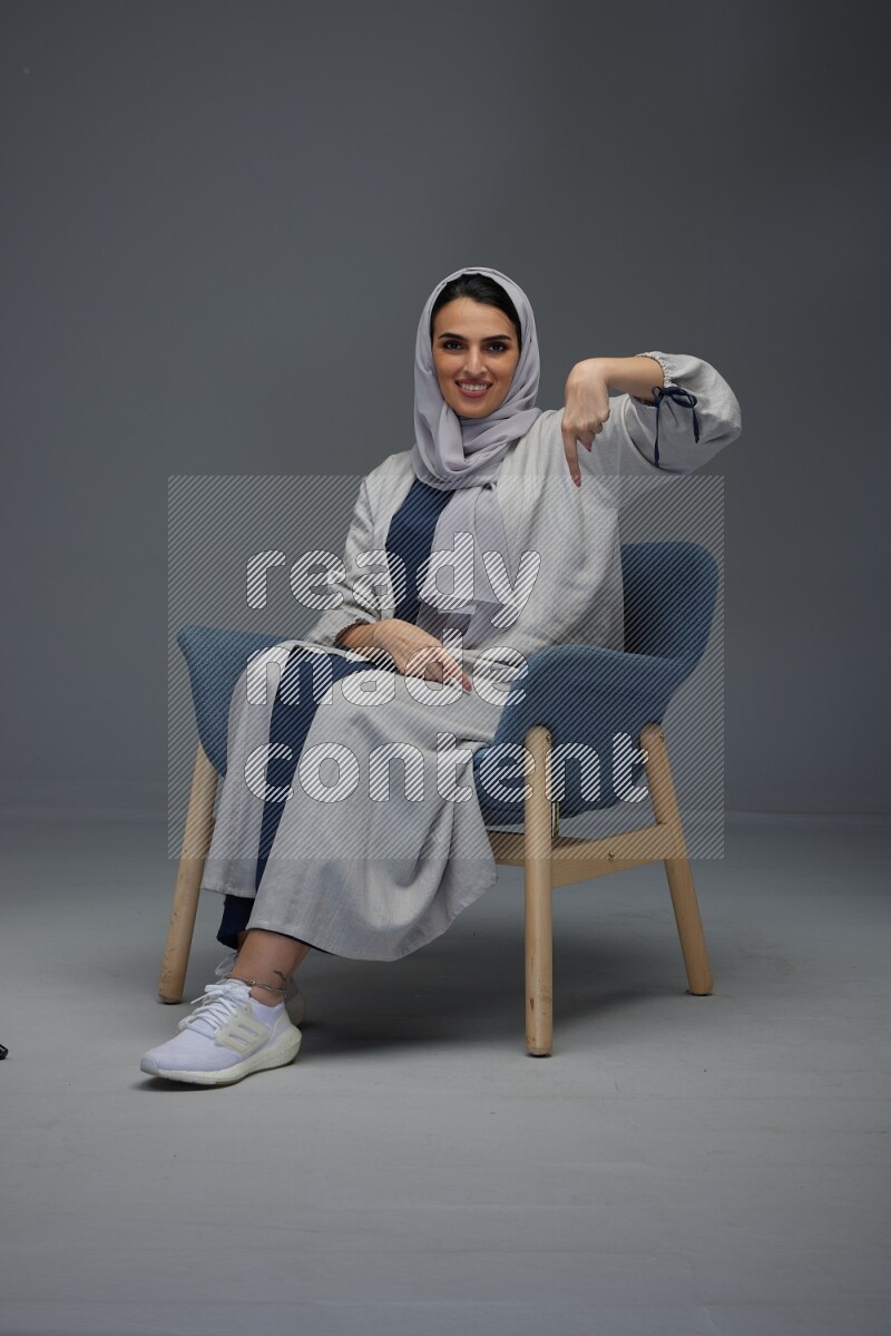 A Saudi woman wearing a light gray Abaya and head scarf sitting on a dark grey chair while making multi hand gestures eye level on a grey background