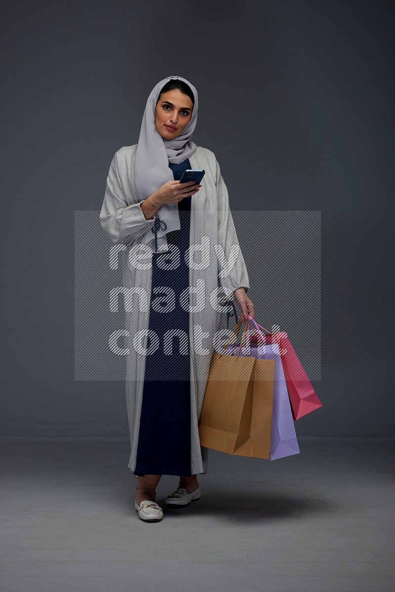 A Saudi woman wearing a light gray Abaya and head scarf standing and holding shopping bags making different poses eye level on a grey background
