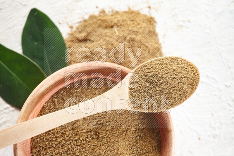 A wooden bowl and wooden spoon full of cumin powder on textured white flooring