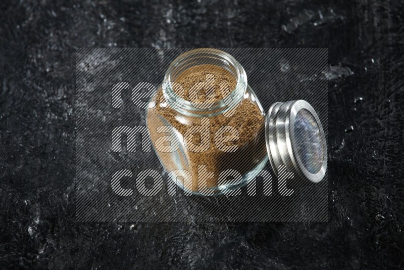 A glass spice jar full of cumin powder on a textured black flooring