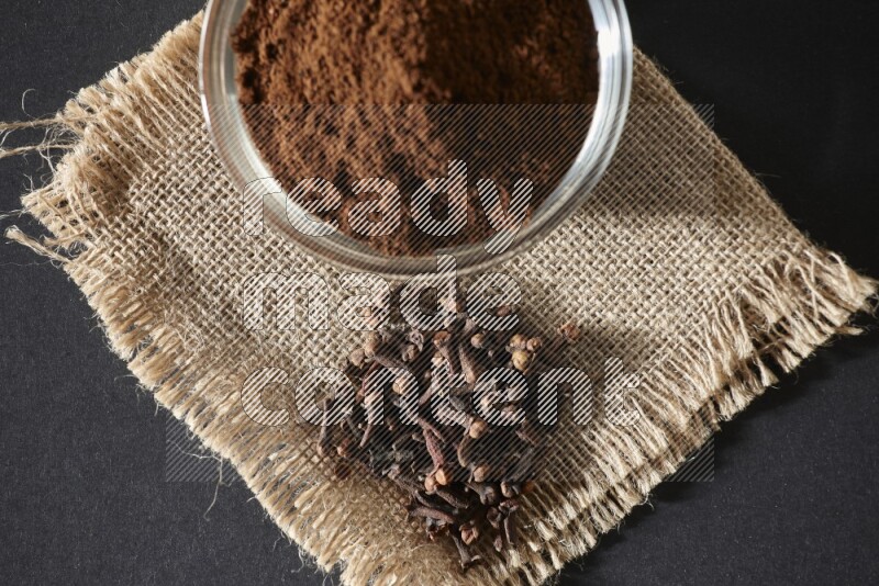 A glass bowl full of cloves powder with cloves grains on a burlap piece on a black flooring