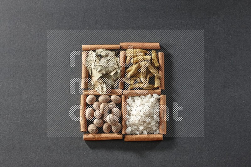 4 مربعات من عيدان القرفة ممتلئة بأوراق الغار والملح الأبيض وأصابع الكركم وجوزة الطيب على أرضية سوداء