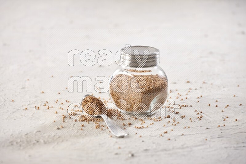 A glass spice jar and a metal spoon full of mustard seeds on a textured white flooring