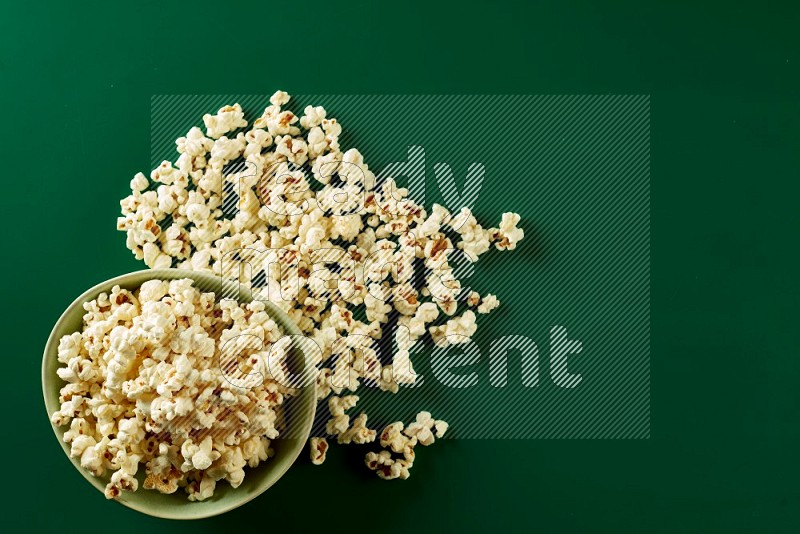 A green ceramic bowl full of popcorn with popcorn beside it on a green background in a top view shot