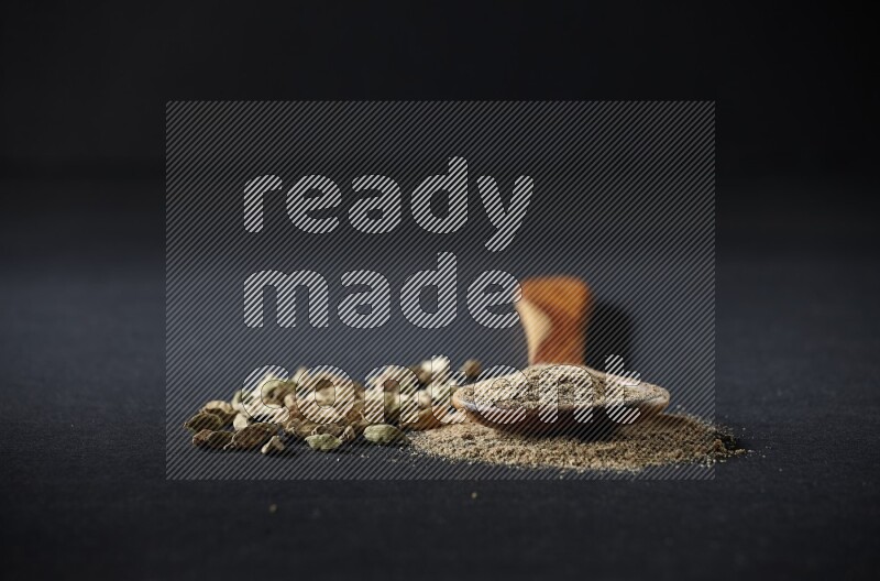A wooden ladle full of cardamom powder and cardamom seeds beside it on black flooring