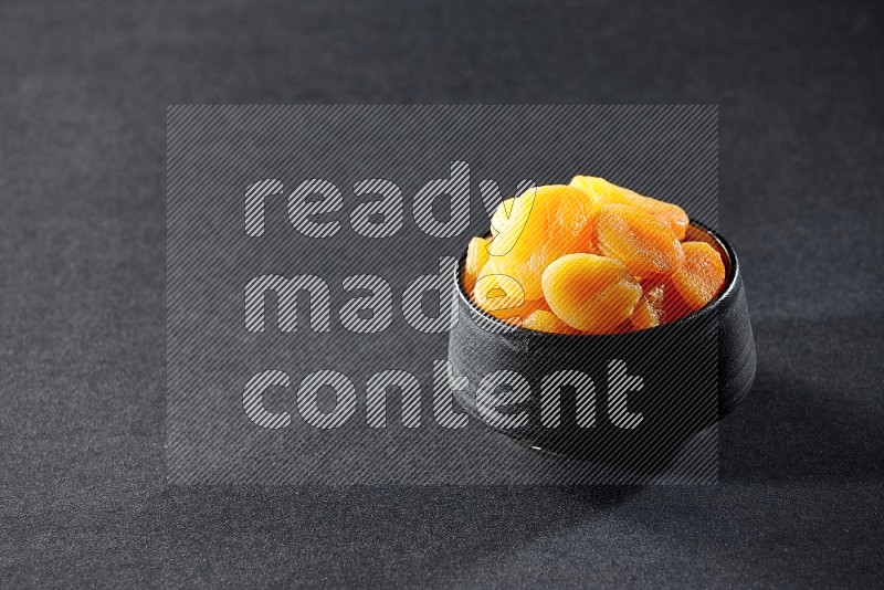 A black ceramic bowl full of dried apricots on a black background in different angles