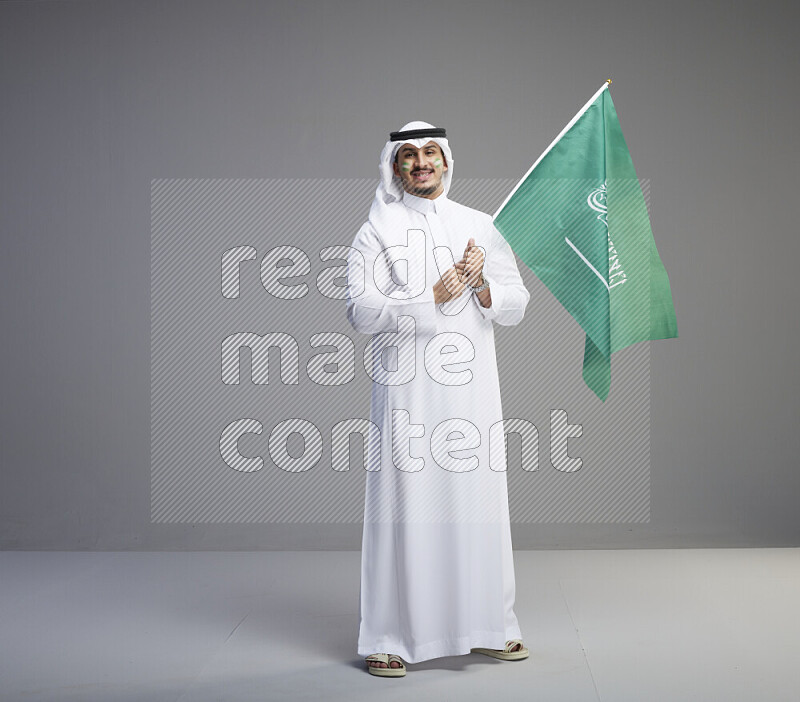 A Saudi man standing wearing thob and white shomag with face painting raising big Saudi flag on gray background