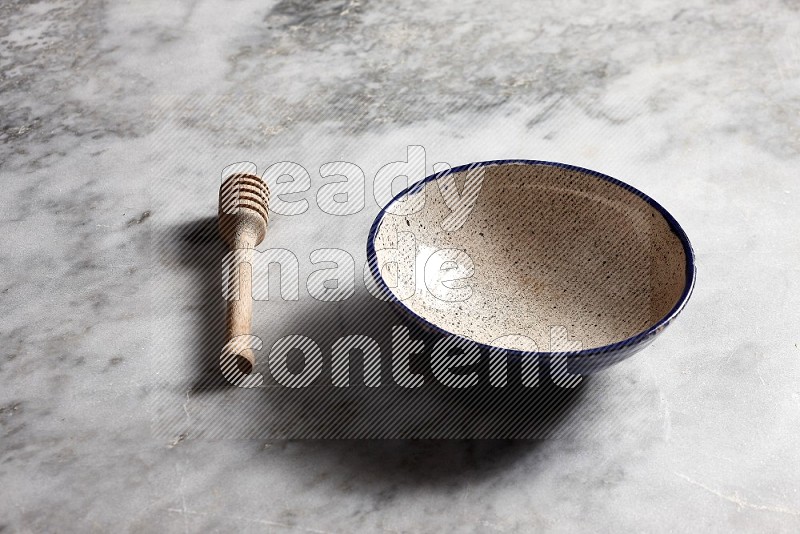 Multicolored Pottery bowl with wooden honey handle on the side with grey marble flooring, 45 degree angle